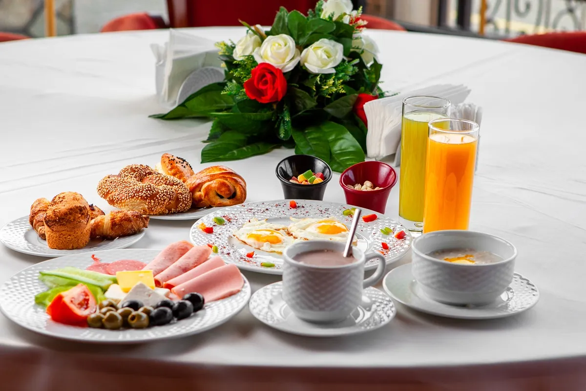 Mesa com pratos de ovos fritos, frios, pães, sucos, café e um arranjo de flores ao centro, representando um café da manhã sofisticado.