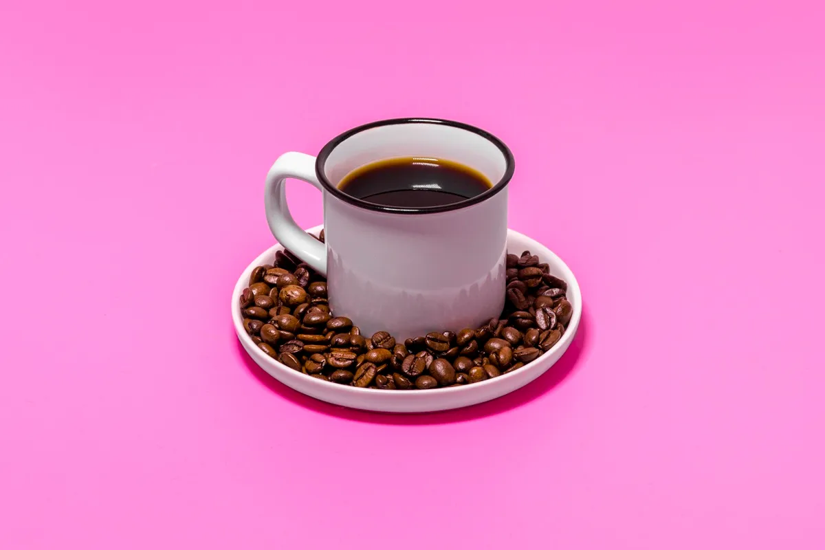 Xícara branca com borda preta cheia de café preto, colocada sobre um prato com grãos de café torrados, em fundo rosa vibrante.