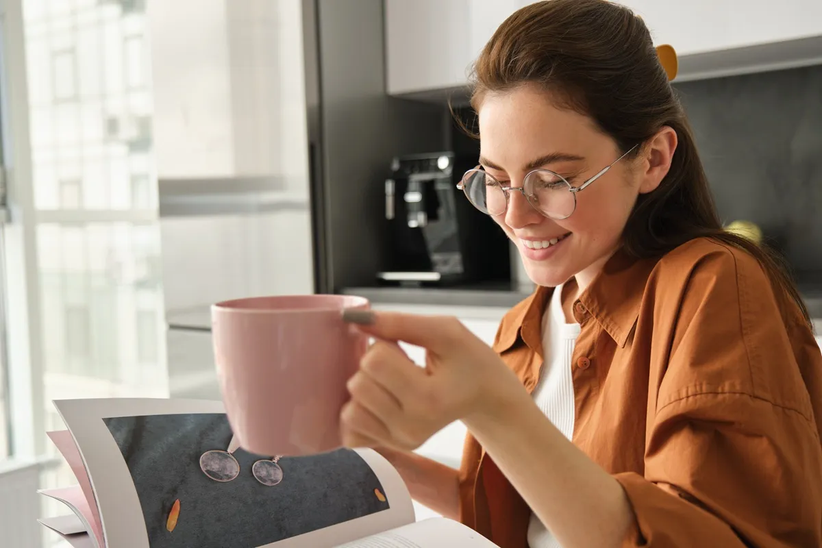 Mulher sorridente segurando uma caneca rosa enquanto lê um livro, em uma cozinha iluminada e moderna.