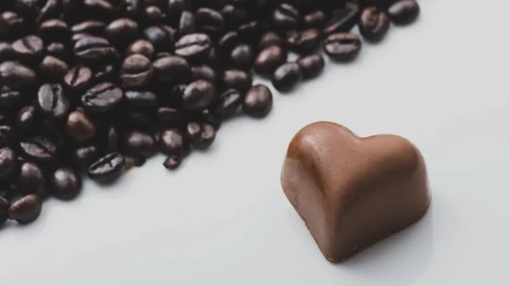 Bombom de chocolate em formato de coração ao lado de grãos de café sobre uma superfície branca.