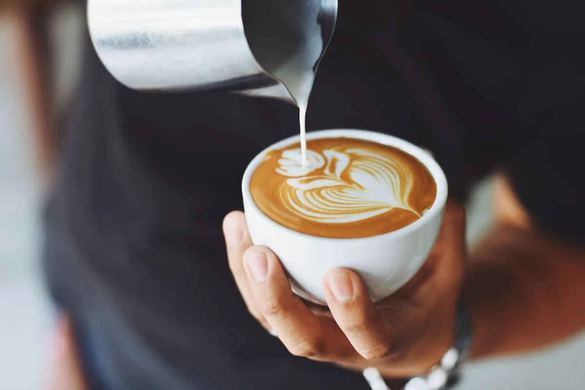 Mãos de uma pessoa derramando leite vaporizado de uma jarra de metal em uma xícara branca, criando um desenho de folha (latte art) sobre o café, ilustrando a palavra-chave "cappuccino caseiro" para uma receita ou tutorial.