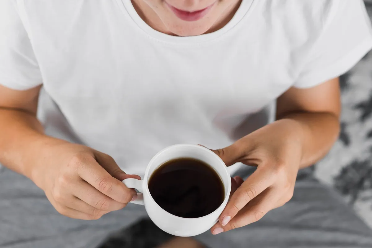 Pessoa segurando uma xícara branca de café preto quente, vestindo uma camiseta branca e calça confortável, aproveitando um momento relaxante.