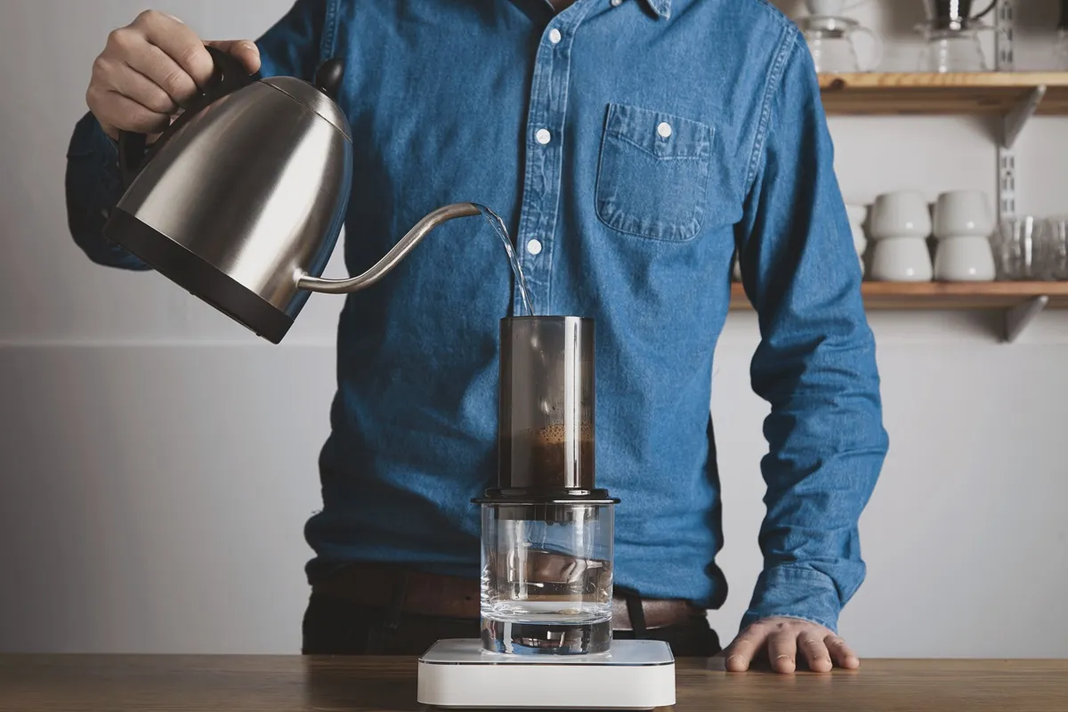 Barista preparando café com Aeropress, despejando água quente de um ebulidor de bico fino sobre o pó de café, com uma balança digital e um copo de vidro para extração.