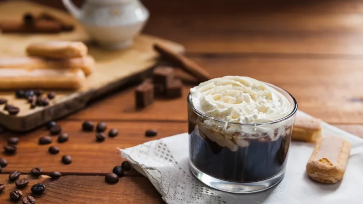 Taça de vidro com gelatina de café coberta por chantilly, acompanhada de biscoitos champanhe sobre uma mesa de madeira rústica.