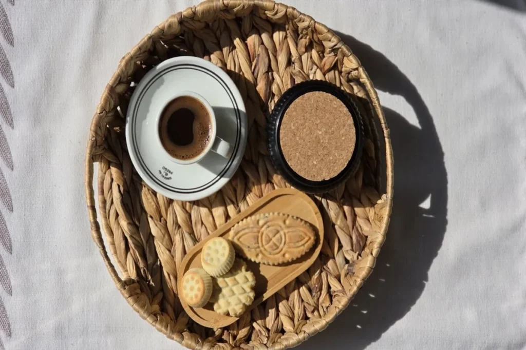 Uma xícara de café, um porta-copos e biscoitos em um prato de madeira, tudo servido em uma bandeja de palha redonda sobre uma toalha de mesa branca, representando opções para um café da manhã para diabéticos.