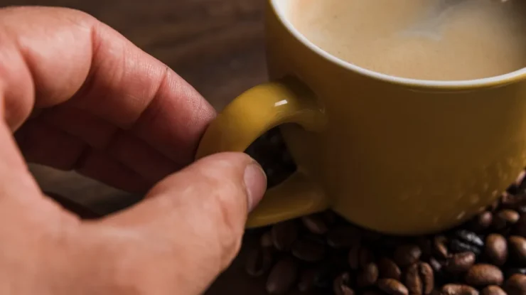 Mão segurando a alça de uma xícara amarela com café cremoso, cercada por grãos de café torrados sobre uma superfície de madeira.