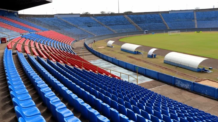 Vista ampla do Estádio do Café, em Londrina, mostrando as arquibancadas vazias com assentos vermelhos e azuis. O campo de futebol ao centro possui gramado bem cuidado e áreas técnicas cobertas. O estádio apresenta estrutura clássica, sendo um importante palco para o futebol paranaense.