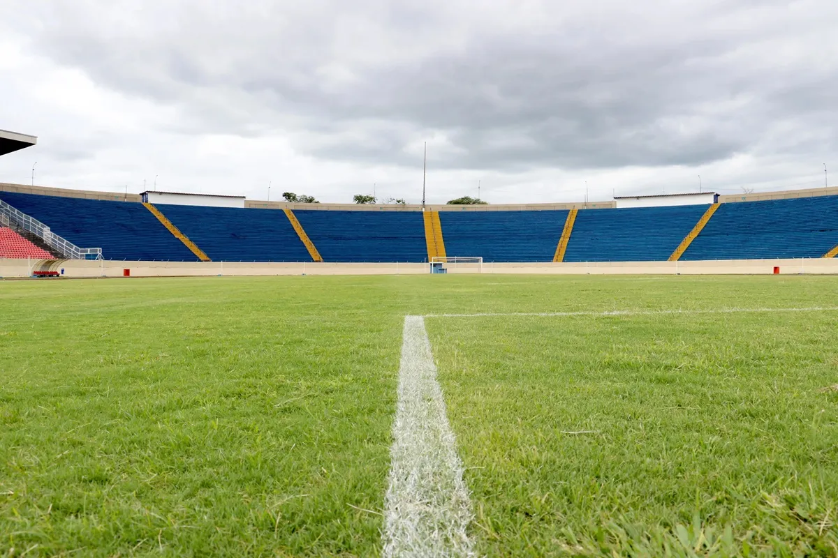 Imagem do Estádio do Café, em Londrina, vista a partir do gramado. O campo verde bem cuidado contrasta com as arquibancadas azuis e amarelas ao fundo. O céu nublado adiciona uma atmosfera clássica ao cenário do estádio.
