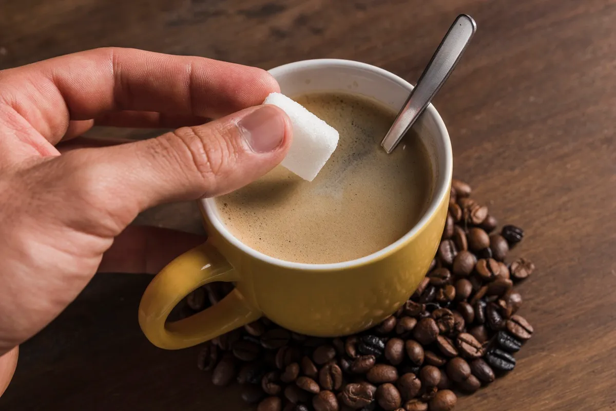 Mão masculina segurando um cubo de açúcar prestes a ser adicionado a uma xícara amarela de café, cercada por grãos de café sobre uma mesa de madeira.