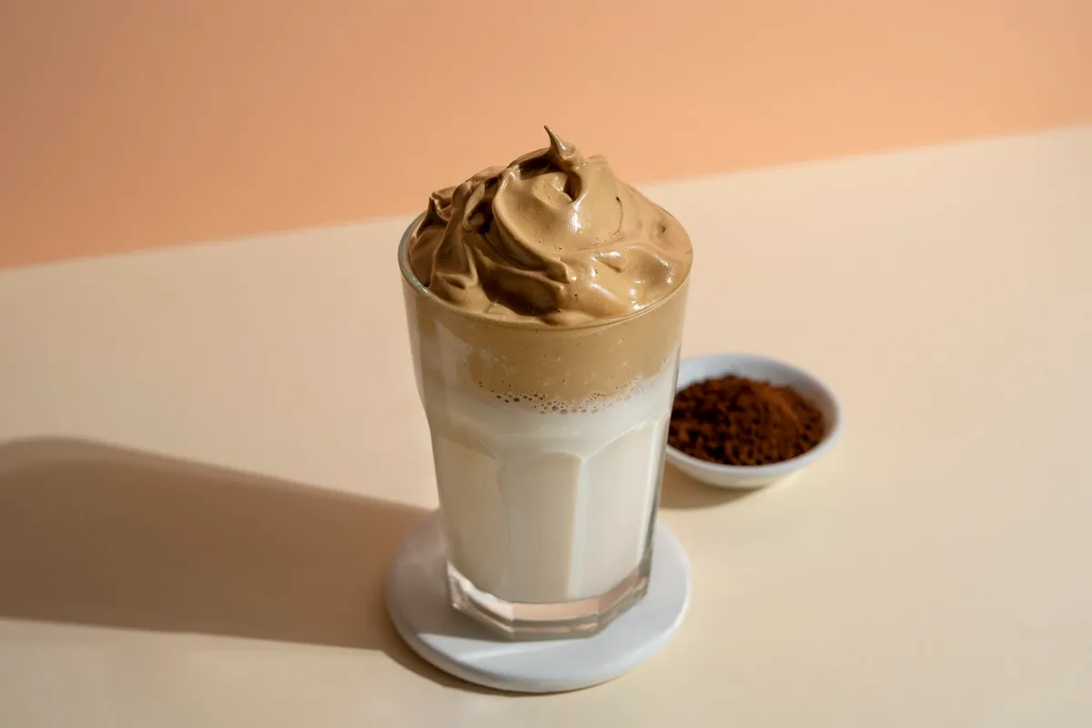 Copo de café Dalgona com espuma cremosa sobre leite gelado, acompanhado de um pratinho com café solúvel, em fundo bege sofisticado.