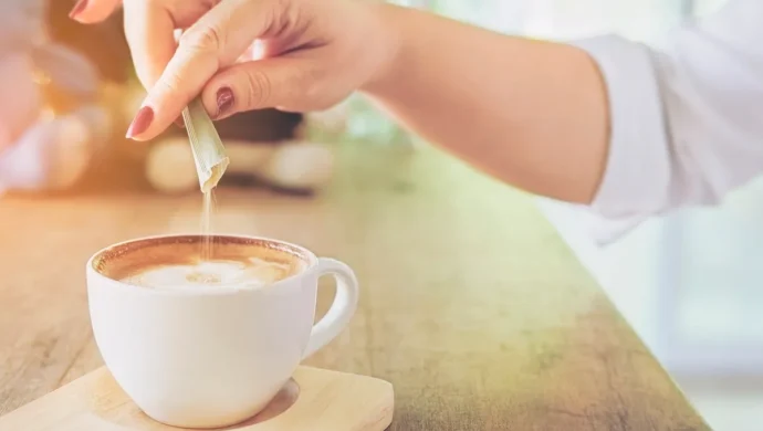 Mão feminina adicionando adoçante em um café cremoso em uma xícara branca sobre uma mesa de madeira, com um ambiente iluminado ao fundo.