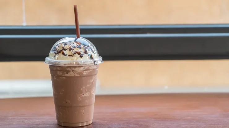 Copo de milkshake de café coberto com chantilly e calda de chocolate, servido com tampa plástica e canudo marrom sobre uma mesa de madeira.
