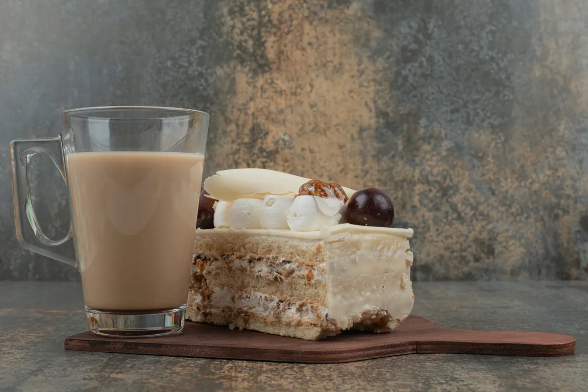 Fatia de pavê de doce de leite com cobertura cremosa, decorada com chocolate branco e confeitos, acompanhada de um café com leite em caneca de vidro.