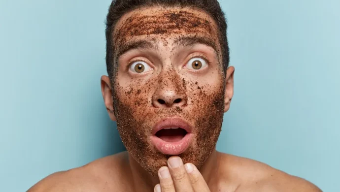 Homem surpreso com máscara esfoliante de café no rosto, expressando espanto enquanto toca o queixo.