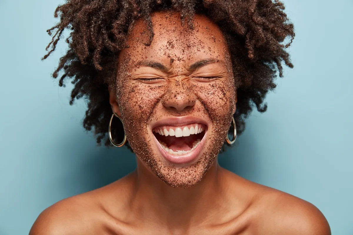 Mulher sorrindo com esfoliante de café no rosto, expressando felicidade e bem-estar com os olhos fechados.