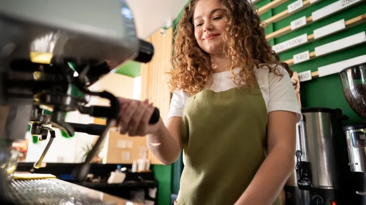 Barista preparando café expresso em máquina profissional, vestindo avental verde, representando o atendimento em uma franquia de café.