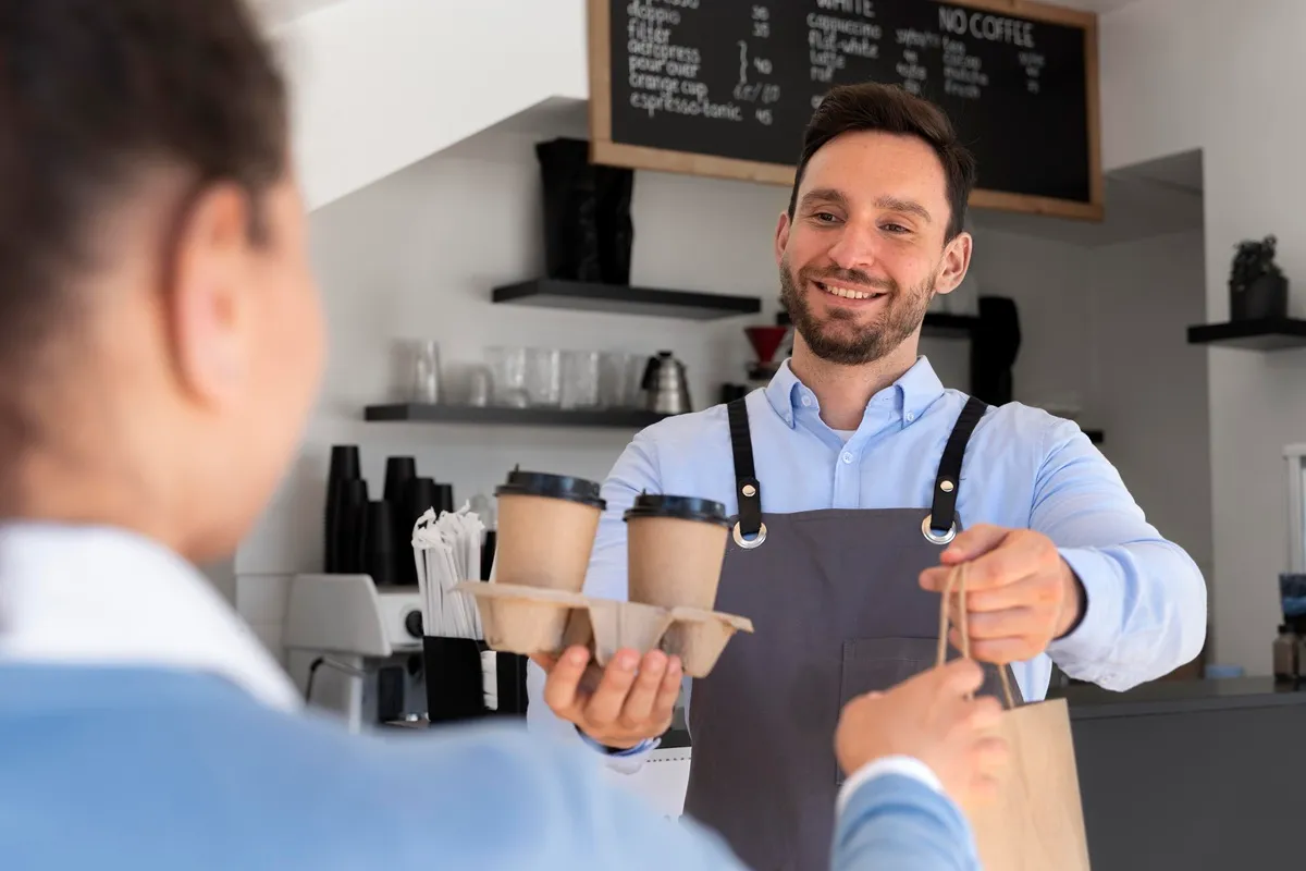 Atendente sorridente entregando pedido com copos de café e sacola para cliente em uma cafeteria moderna.