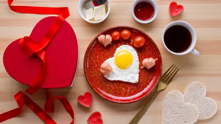 Imagem de um café da manhã romântico com ovo frito em formato de coração, tomates, salsichas, pão, café e uma caixa de presente com fita vermelha