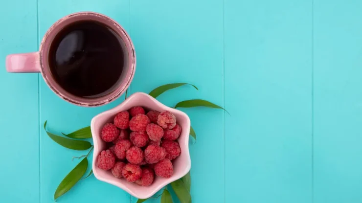 Xícara de chá quente ao lado de tigela com framboesas frescas sobre fundo azul com folhas decorativas.