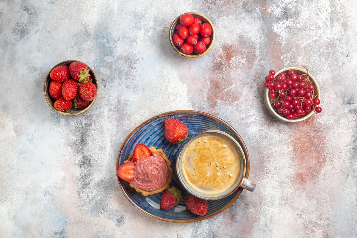 Xícara de café com espuma servida ao lado de cupcake com morango, acompanhada por potes de frutas vermelhas como morangos, groselhas e acerolas, sobre fundo rústico.