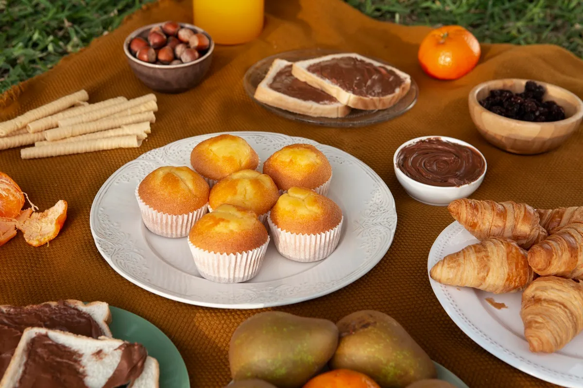 Imagem de um café colonial ao ar livre, com muffins, pães, frutas, torradas com chocolate, sucos e uma atmosfera acolhedora