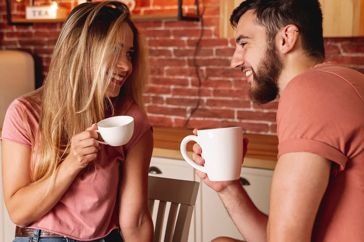 Imagem de um casal sorrindo, segurando xícaras de café enquanto se olham em um ambiente acolhedor de cozinha