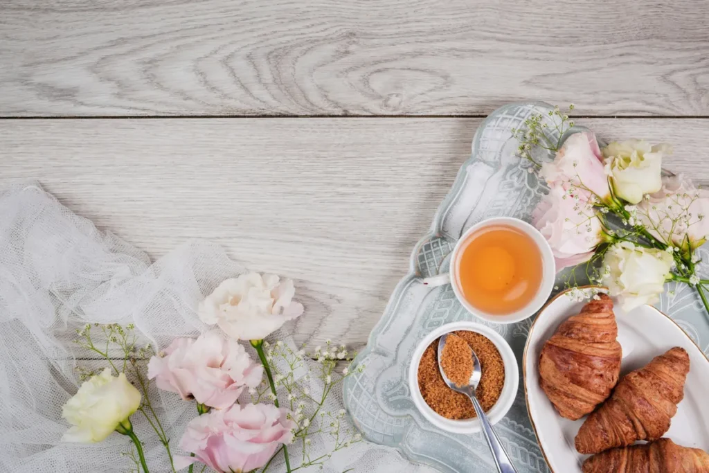 Mesa posta café da manhã com xícara de chá, tigela de açúcar mascavo, croissants e arranjo de flores delicadas sobre fundo de madeira clara.