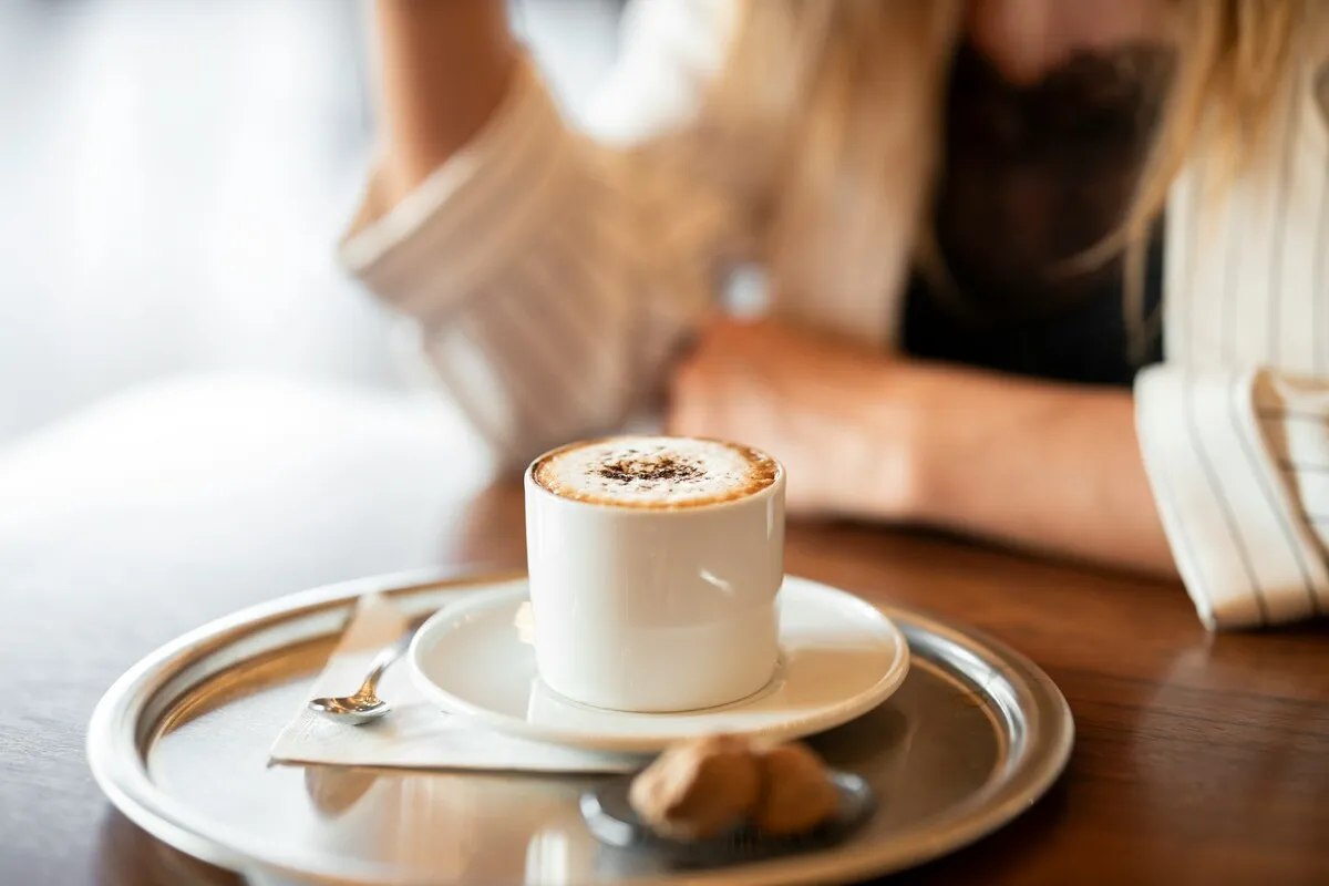 Close de uma xícara de café com espuma servida em bandeja de prata ao lado de uma mulher em cafeteria