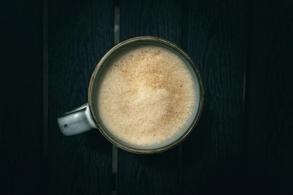 Xícara de cerâmica com café com leite em pó e espuma cremosa, vista de cima, sobre uma superfície escura e texturizada, destacando a textura da bebida.