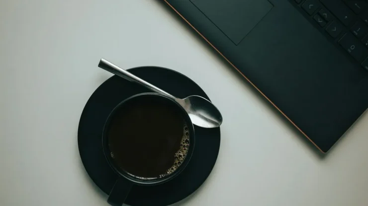 Vista superior de uma xícara de café preto em pires preto com colher de inox ao lado de um notebook sobre mesa branca