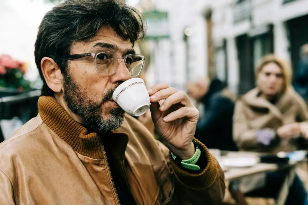 Homem maduro com óculos tomando café em xícara pequena em um café ao ar livre.