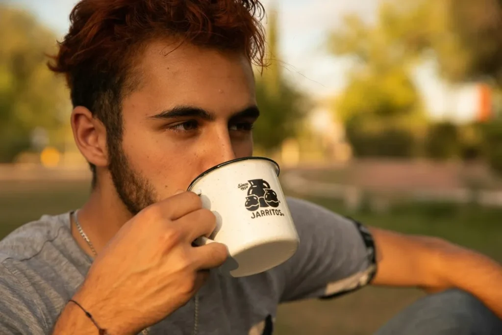 Homem jovem bebendo em uma caneca branca com o logotipo Jarritos, ao ar livre, em um parque.