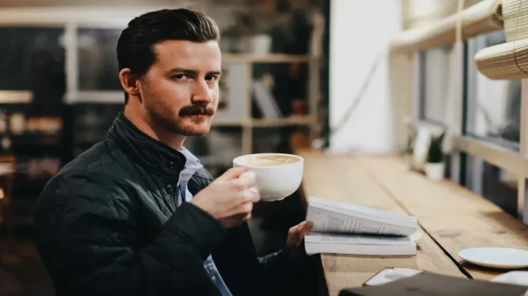 Homem segurando uma xícara de café enquanto lê um livro em cafeteria aconchegante.