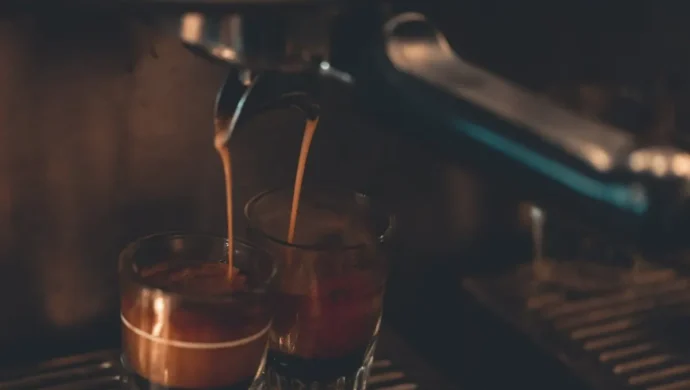 Café espresso sendo preparado em máquina profissional, com duas doses sendo servidas em copos de vidro sob baixa iluminação.