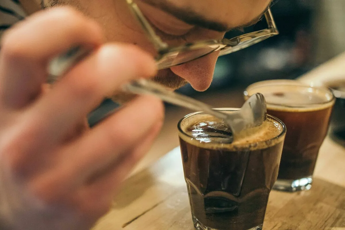 Homem aproximando o rosto de um copo de vidro com café para sentir o aroma, usando colher durante a técnica de cupping