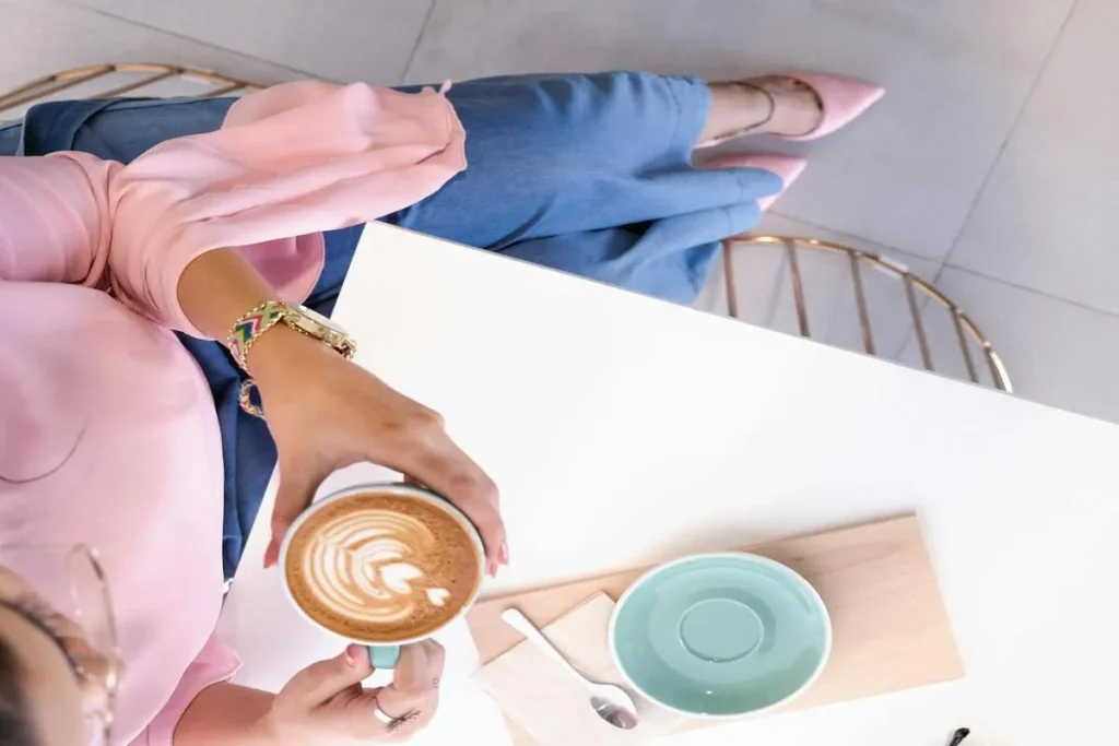 Uma pessoa segura uma chávena de café com leite decorado com a arte de um coração, vista de cima, numa mesa de café moderna. A imagem representa o "coffee break" como um momento de relaxamento e prazer.