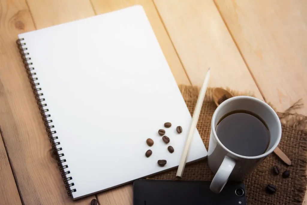 Uma vista de cima de uma mesa de madeira com uma caneca de café, um caderno em branco, um lápis, grãos de café e um smartphone, representando o conceito de um "coffee break".