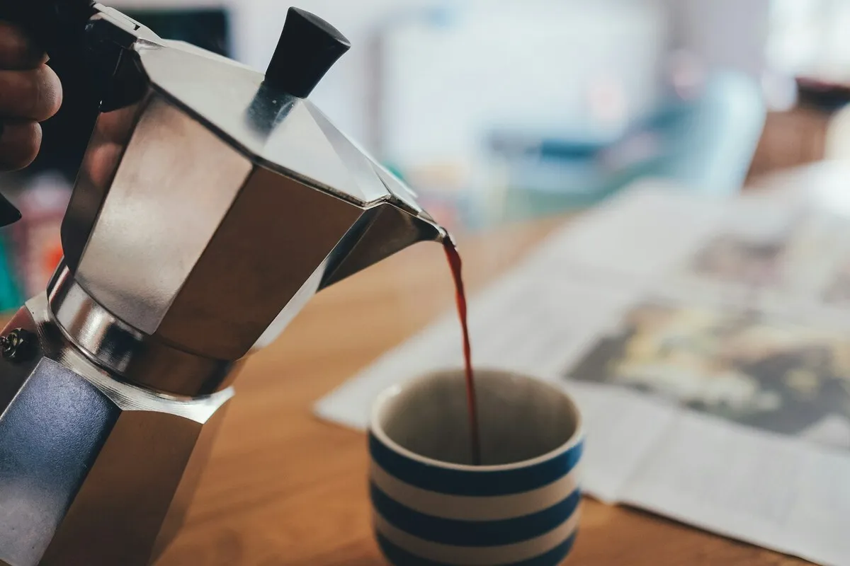 Café sendo servido de uma cafeteira italiana em uma xícara listrada sobre uma mesa com jornal.