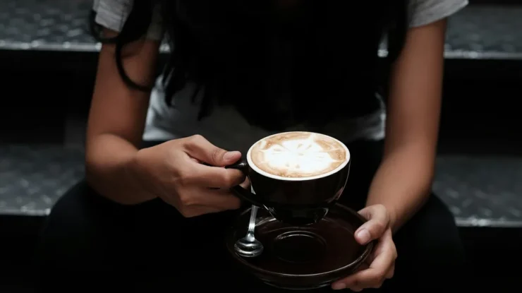 Pessoa segurando xícara de cappuccino com arte no leite, sentada em escadas.