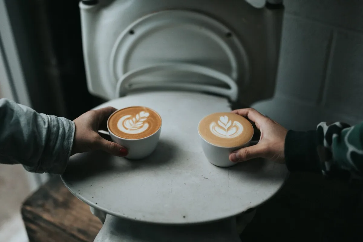 Duas pessoas segurando xícaras de cappuccino com arte no leite sobre uma mesa redonda branca.