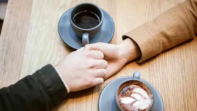 Casal segurando as mãos sobre a mesa com duas xícaras de café em um ambiente acolhedor.