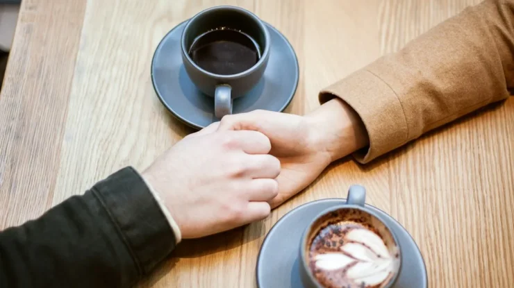 Casal segurando as mãos sobre a mesa com duas xícaras de café em um ambiente acolhedor.