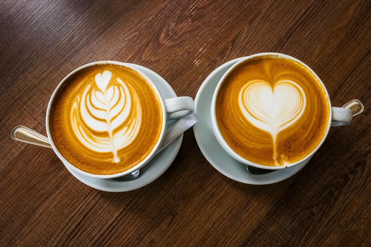 Duas xícaras de cappuccino com arte no leite em formato de coração e folha sobre mesa de madeira.