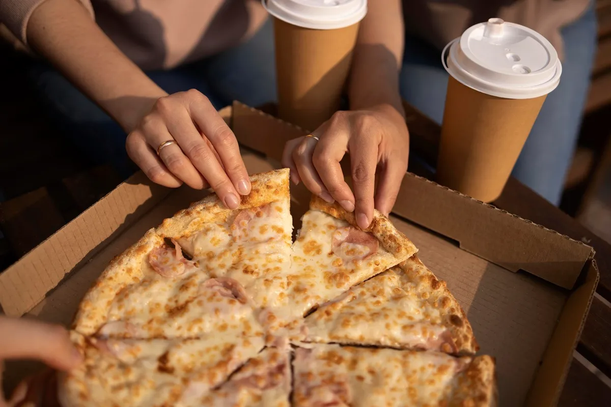 Duas pessoas pegando fatias de pizza de uma caixa de entrega, acompanhadas por copos de café, em um ambiente ao ar livre.