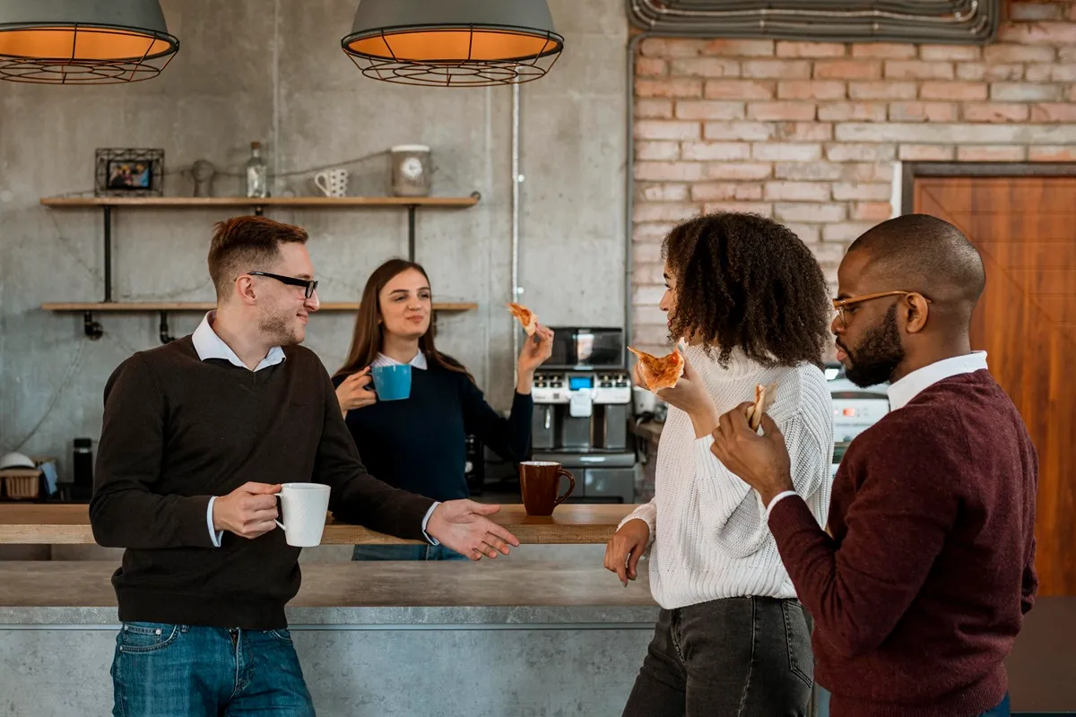 Grupo de colegas de trabalho se socializando no escritório, com café e pizza, em um ambiente moderno e descontraído.