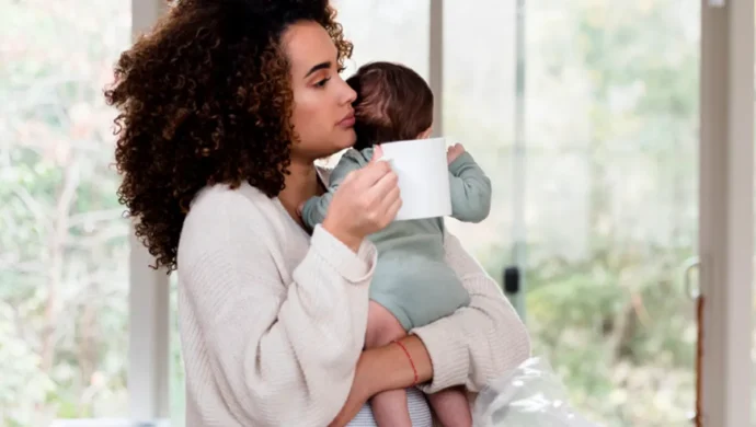 Mãe segurando bebê no colo enquanto toma café em casa, perto da janela.