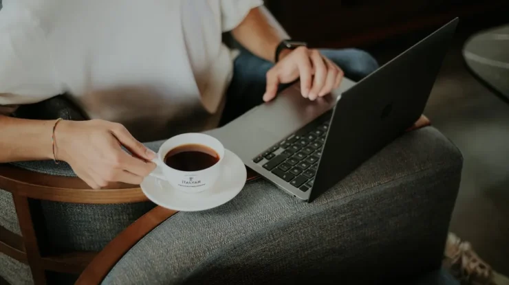 Pessoa usando notebook em uma poltrona confortável enquanto segura uma xícara de café com pires, ambiente acolhedor e moderno.