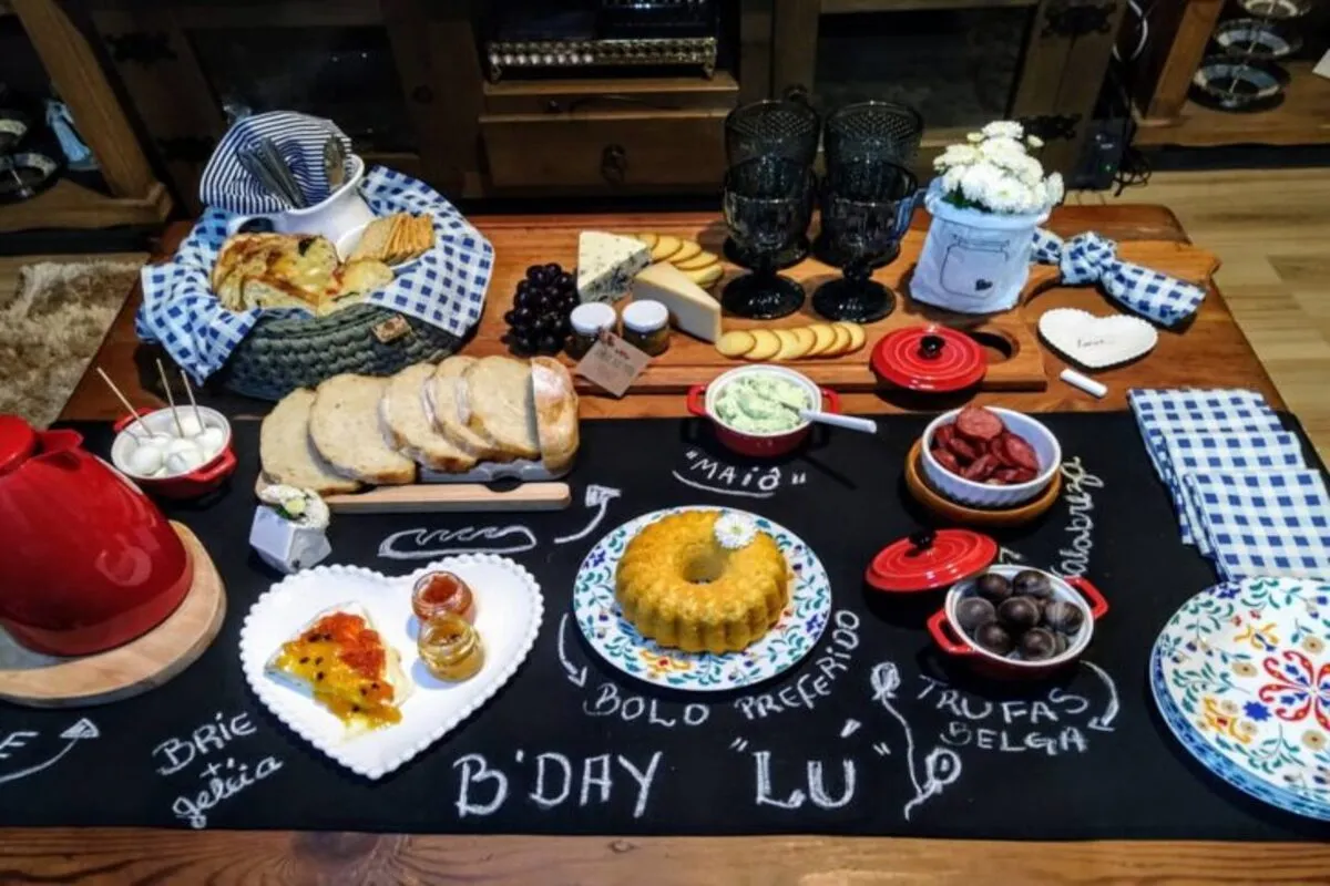 Mesa de café da manhã de aniversário ricamente decorada com pães artesanais, queijos, bolo, trufas e a mensagem "B'DAY 'LU'".