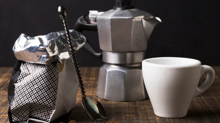 Pacote de café aberto, grãos de café espalhados, uma colher vintage, uma cafeteira italiana e uma xícara branca sobre uma superfície de madeira escura, ilustrando a preparação de "café para cafeteira italiana".