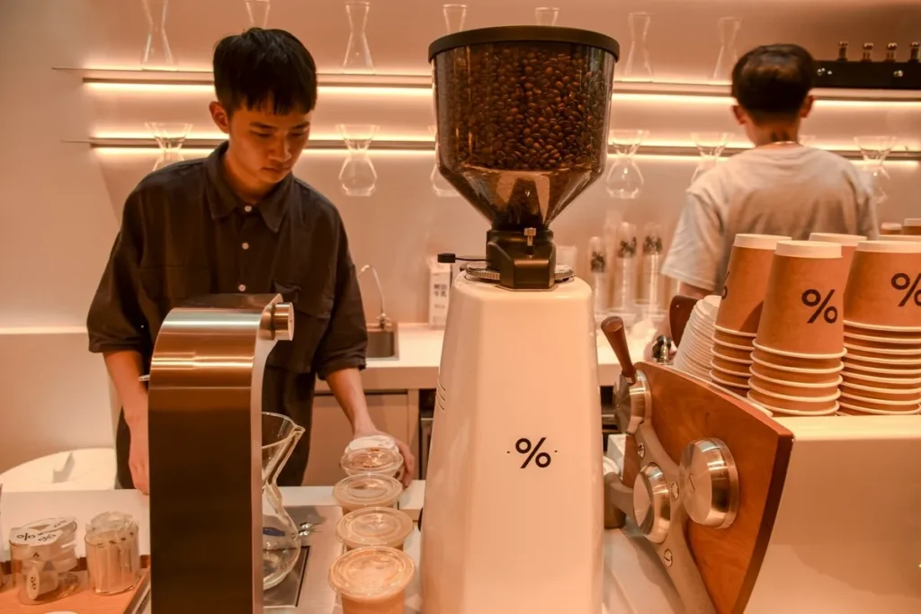 Barista trabalhando em uma cafeteria moderna com uma cafeteira que moe o café na hora, mostrando grãos de café inteiros no moedor e copos de papel prontos para servir.